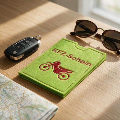 Fahrzeugscheinhülle aus hellgrünem Filz mit rot gesticktem Schriftzug KFZ-Schein und Motorrad auf Holztisch neben Autoschlüssel, Sonnenbrille und Straßenkarte