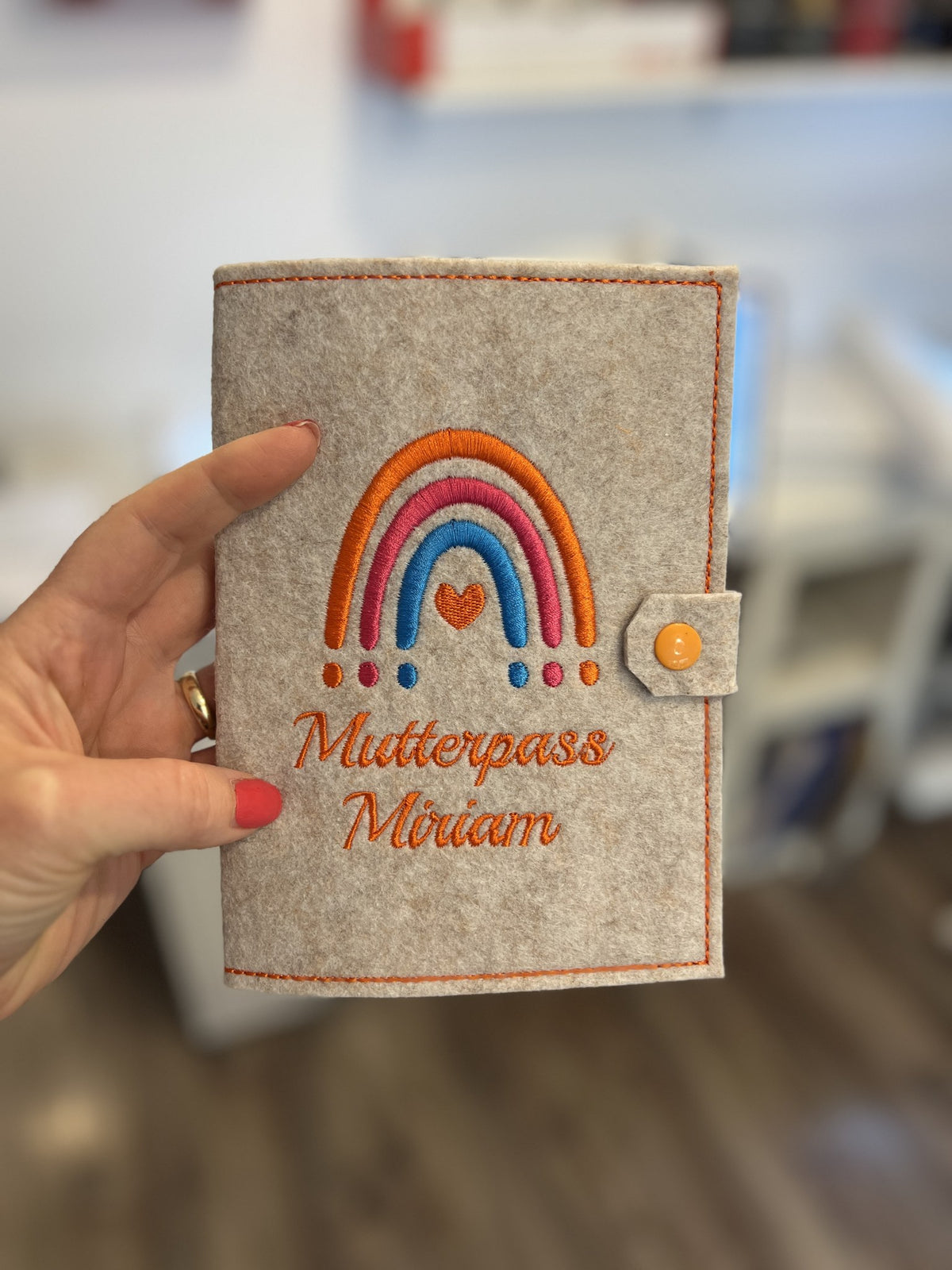 Hand hält Mutterpasshülle aus Filz mit Regenbogen und Name Miriam, Druckknopf in Orange
