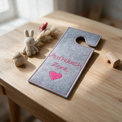 Türanhänger aus grauem Filz mit gesticktem Text „Aufhübsch Zone“ und Herz auf Holztisch neben Stoffhase und Trockenblumen